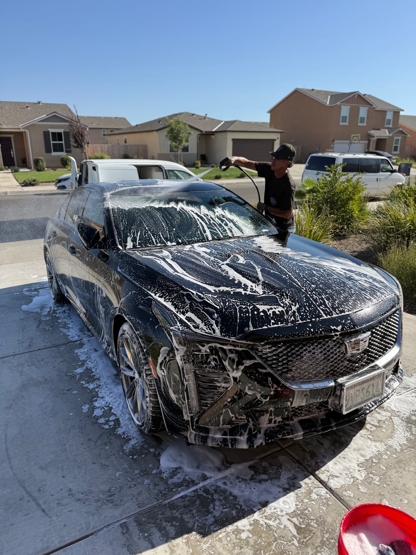 Black Cadillac covered in thick snow foam being hand-washed by a mobile detailer in a driveway